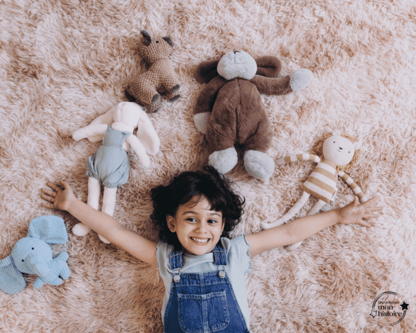 Jeune fille souriante, écartant les bras, allongée sur une moquette beige à poil long. Au dessus et autour d'elle, sont disposés plusieurs doudous.